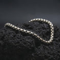 Load image into Gallery viewer, Polished silver box chain bracelet arranged across dark volcanic stone, highlighting the clean square-link structure and smooth reflective finish.