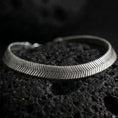 Load image into Gallery viewer, Oxidized sterling silver herringbone bracelet displayed on black volcanic rock, showing its slim flat profile and chevron texture.