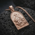 Load image into Gallery viewer, The rose gold plated INRI pendant is arranged on rugged volcanic rock, emphasizing the sculpted figure of Jesus, the cross above him, and the ornamental arched frame in a refined macro image.