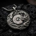 Load image into Gallery viewer, An oxidized dragon and phoenix pendant rests on dark volcanic rock, showing the central Yin Yang symbol, octagonal Bagua design, detailed scales, flowing feathers, and cloud motifs in a macro studio close-up.
