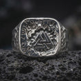Load image into Gallery viewer, The polished silver Viking signet ring is displayed on textured black volcanic rock, highlighting the central Valknut emblem, rune border, sculpted skull, and detailed masculine shoulders in a premium macro photograph.