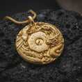 Load image into Gallery viewer, A gold plated Chinese dragon and phoenix pendant lies on rugged volcanic rock, featuring a central Yin Yang emblem, Bagua-inspired lines, layered scales, ornamental feathers, and symbolic cloud details in a luxury macro close-up.