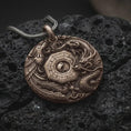 Load image into Gallery viewer, Bronze dragon and phoenix pendant rests on dark volcanic rock, showing the central Yin Yang symbol, octagonal Bagua design, detailed scales, flowing feathers, and cloud motifs in a macro studio close-up.