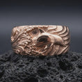 Load image into Gallery viewer, The rose gold plated ocean wave ring is arranged on black volcanic rock, emphasizing the raised dolphin motif, curling wave patterns, and substantial unisex band in a refined macro image.