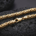 Load image into Gallery viewer, A gold plated Byzantine chain necklace lies across rugged volcanic rock, featuring bold interlocking links, a lobster clasp, and a substantial masculine chain profile in a luxury macro studio close-up.
