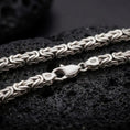 Load image into Gallery viewer, The polished silver Byzantine chain necklace is displayed on textured black volcanic rock, highlighting the rounded woven link pattern, secure clasp, and 925 silver markings in a premium macro photograph.