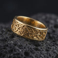 Load image into Gallery viewer, A gold plated Celtic knot band rests on rugged volcanic rock, featuring raised interwoven details and a symbolic continuous pattern across the wide ring in a luxury macro studio close-up.