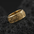 Load image into Gallery viewer, A gold plated floral spinner ring rests on rugged volcanic rock, featuring raised flower details, geometric accents, and an ornamental wide band design in a luxury macro studio close-up.