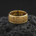 Load image into Gallery viewer, A gold plated Irish spinner ring sits on textured volcanic rock, showcasing the woven Celtic knot motif, symbolic band pattern, and elegant wide profile in a luxury macro shot.