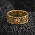 Load image into Gallery viewer, A gold plated Aztec spinner ring rests on rugged volcanic rock, featuring symbolic panel engravings, stepped linework, and a bold unisex wedding band profile in a luxury macro studio close-up.
