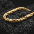 Load image into Gallery viewer, A gold plated Byzantine chain necklace lies on rugged volcanic rock, featuring bold 6mm interlocking links and a substantial masculine profile in a luxury macro studio close-up.