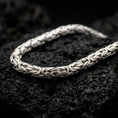 Load image into Gallery viewer, The polished silver 6mm Byzantine chain necklace is displayed across textured black volcanic rock, highlighting its dimensional round links and intricate woven construction in a premium macro photograph.