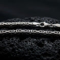 Load image into Gallery viewer, An oxidized 4mm square Byzantine chain necklace rests on dark volcanic rock, highlighting its tightly woven interlocking links and bold sterling silver structure in a macro studio close-up.