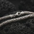 Load image into Gallery viewer, The polished silver 4mm round Byzantine chain necklace is displayed on textured black volcanic rock, highlighting the woven link pattern, lobster clasp, and 925 silver stamp in a premium macro photograph.