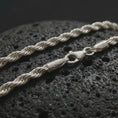 Load image into Gallery viewer, The polished silver 3mm rope chain necklace is displayed on textured black volcanic rock, highlighting the braided twist pattern, secure clasp, and refined everyday profile in a premium macro photograph.