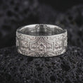 Load image into Gallery viewer, The polished silver ornate cross ring is displayed on textured black volcanic rock, highlighting its symmetrical ornamental design, raised silver details, smooth rounded edges, and wide band profile in a premium macro photograph.