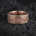 Load image into Gallery viewer, The rose gold plated ornate cross band ring is arranged on black volcanic rock, emphasizing its symmetrical ornamental relief, floral centerpiece, decorative cross motifs, and wide medieval-inspired band design.