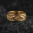 Load image into Gallery viewer, A gold plated floral silver ring rests on rugged volcanic rock, showcasing raised flowers, botanical foliage, dark recessed detailing, and an ornate wide band profile in a luxury macro studio close-up.