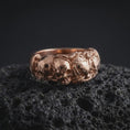 Load image into Gallery viewer, The rose gold plated skull and rose band ring is arranged on black volcanic rock, emphasizing its detailed skull centerpiece, floral relief, organic foliage, and dark romantic gothic-inspired design.