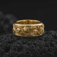 Load image into Gallery viewer, A gold plated skull snake ring rests on rugged volcanic rock, showcasing raised skull motifs, a coiling serpent, detailed roses, and dramatic memento mori-inspired relief in a luxury macro studio close-up.