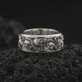 Load image into Gallery viewer, The polished silver gothic skull and snake ring is displayed on textured black volcanic rock, highlighting the three-dimensional skulls, serpent scales, floral roses, and substantial wide band profile in a premium macro photograph.