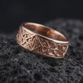 Load image into Gallery viewer, The rose gold plated Celtic knot ring is arranged on black volcanic rock, emphasizing the engraved interlace design, dimensional band texture, and historical-inspired craftsmanship in a refined macro image.