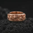 Load image into Gallery viewer, The rose gold plated skull and serpent band is arranged on black volcanic rock, emphasizing its sculpted skulls, winding snake, floral rose details, and bold dark romantic jewelry design in a detailed macro image.