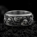 Load image into Gallery viewer, An oxidized sterling silver skull snake rose ring rests on dark volcanic rock, featuring sculpted skulls, a winding serpent, raised roses, and deep recessed detailing in a dramatic macro studio close-up.