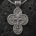 Load image into Gallery viewer, An oxidized sterling silver double sided Saint Michael cross pendant rests on dark volcanic rock, showing a detailed crucifixion scene with sacred figures, radiant motifs, and bold relief in a dramatic macro studio close-up.