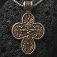 Load image into Gallery viewer, Bronze sterling silver double sided Saint Michael cross pendant rests on dark volcanic rock, showing a detailed crucifixion scene with sacred figures, radiant motifs, and bold relief in a dramatic macro studio close-up.