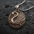 Load image into Gallery viewer, A bronze sterling silver swan pendant rests on dark volcanic rock, featuring a graceful curved neck, layered feather wings, flowing water scrollwork, and dark recessed details in a premium macro close-up.