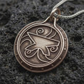 Load image into Gallery viewer, A bronze sterling silver Zenithar-inspired amulet pendant rests on dark volcanic rock, featuring a raised geometric merchant symbol, spiral details, dotted border, and dark recessed circular background.