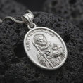 Load image into Gallery viewer, Polished sterling silver Saint Homobonus pendant displayed on dark volcanic stone, highlighting the saint portrait, halo, staff, religious details, and oval medal frame.