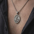 Load image into Gallery viewer, Oxidized sterling silver Saint Homobonus necklace worn on the chest, showing the detailed Catholic saint medal, halo, staff, and SAINT HOMOBONUS inscription.