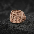 Load image into Gallery viewer, Rose gold plated Calvary ring displayed on textured volcanic stone, showcasing Jesus on the central cross, two crucified thieves, architectural details, and an ornate Christian signet profile.