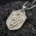 Load image into Gallery viewer, Polished sterling silver dragon shield necklace displayed on dark volcanic stone, highlighting the sculpted wings, scaled coiled body, fierce dragon head, and heraldic shield frame.