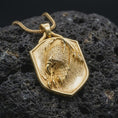 Load image into Gallery viewer, Gold plated eagle shield pendant resting on black volcanic rock, featuring a powerful eagle head, detailed plumage, sharp claws, and bold heraldic medallion design.
