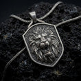 Load image into Gallery viewer, Oxidized sterling silver roaring lion shield pendant on black volcanic rock, featuring a fierce lion head, open jaws, sharp fangs, flowing mane, and textured heraldic shield background.