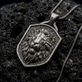 Load image into Gallery viewer, Oxidized sterling silver regal lion shield pendant on black volcanic rock, featuring a majestic lion portrait, calm expression, flowing mane, and textured heraldic shield background.