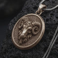 Load image into Gallery viewer, Bronze Aries ram pendant on black volcanic rock, featuring a sculpted ram head, large curled horns, textured background, and oval medallion frame.