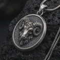 Load image into Gallery viewer, Oxidized sterling silver Aries ram pendant on black volcanic rock, featuring a sculpted ram head, large curled horns, textured background, and oval medallion frame.