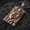 Load image into Gallery viewer, Bronze Gemini twins pendant on black volcanic rock, featuring two sculpted faces resting together, detailed facial expressions, and an intricate geometric rectangular frame.
