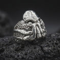 Load image into Gallery viewer, Polished sterling silver Lovecraft Cthulhu ring displayed on dark volcanic stone, highlighting the massive sculpted forehead, sinister eye sockets, curling tentacles, and intricate circular sucker patterns wrapping around the band.