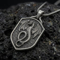 Load image into Gallery viewer, Oxidized sterling silver coiled dragon shield pendant on black volcanic rock, featuring a winged dragon, detailed scales, curled serpentine body, and textured medieval shield.