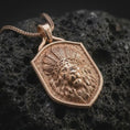 Load image into Gallery viewer, Rose gold plated warrior lion pendant displayed on dark volcanic stone, showcasing the majestic lion face, intricately carved feather crown, textured recessed background, and decorative shield-shaped frame.