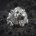 Load image into Gallery viewer, Polished silver tiger head ring on dark lava stone, showcasing a fierce roaring expression, deep-set eyes, pronounced nose, textured mane-like fur, and statement ring silhouette.