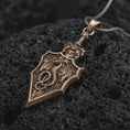 Load image into Gallery viewer, Bronze dragon sword pendant displayed on textured black volcanic rock, featuring a winged dragon coiled around a medieval-style sword blade, intricate scales, decorative hilt, angular edges, and dark oxidized silver finish.