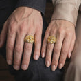 Load image into Gallery viewer, Gold plated Masonic skull ring worn on a finger, highlighting the dimensional skull face, raised Freemason Square and Compasses emblem, flowing eagle feather details, and distinctive biker-inspired jewelry design.