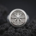 Load image into Gallery viewer, Polished sterling silver nautical compass ring displayed on dark volcanic stone, highlighting the dimensional eight-point star, cardinal direction letters, concentric compass circles, and substantial round signet face.