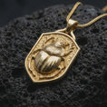 Load image into Gallery viewer, Gold plated stag beetle pendant resting on black volcanic rock, featuring a sculpted insect with prominent antler-like jaws, rounded armored wing covers, and detailed legs against a medieval-style shield.